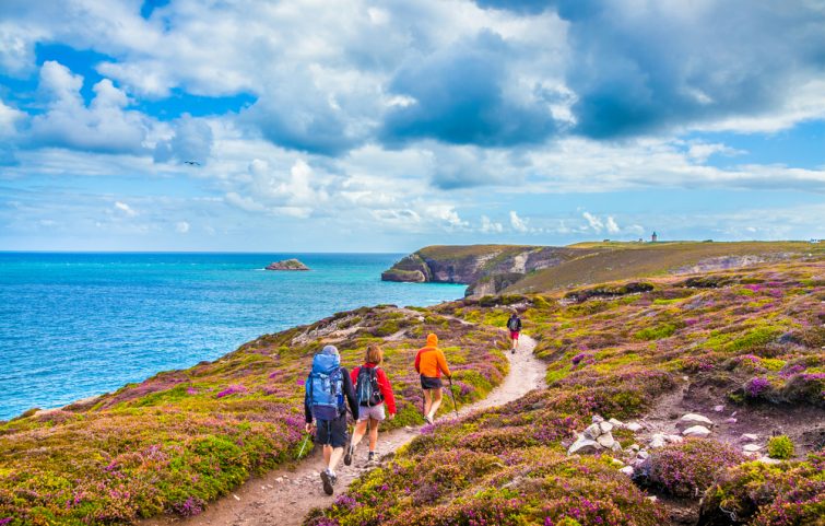 Touristes randonnant le long des belles côtes de Bretagne au célèbre point de vue de la péninsule du Cap Frehel avec phare traditionnel en arrière-plan par jour ensoleillé avec ciel bleu et nuages en été, France