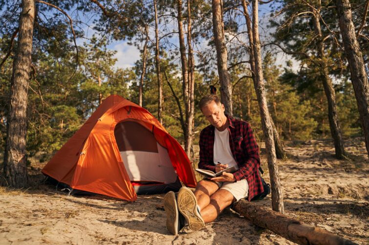 Homme mûr assis sur un tronc près d'un lac tout en écrivant dans un journal lors d'un camping